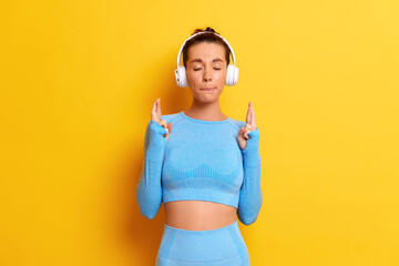 Photo of slender athletic girl in blue yoga suit standing with closed eyes and fingers up, posing on yellow background with earphones, sport lifestyle concept, copy space, high quality photo