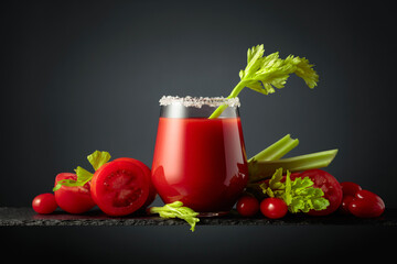 Fresh tomato juice in a glass with a celery stick and a heap of ripe vegetables on a black background.