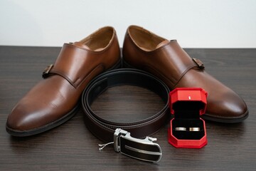 Pair of brown leather shoes, a belt, and wedding rings on a wooden table.
