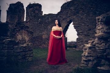Young beautiful female in a medieval red dress posing near ancient ruins