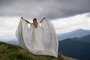Fototapeta na wymiar Woman in a white bridal cape standing atop a hill with a scenic landscape.