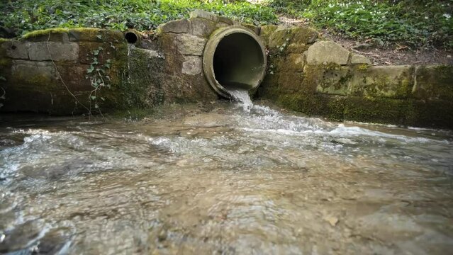 Small water stream running out of an industrial-size concrete pipe in a forest in Europe. Daytime, slow motion, no people, wide angle