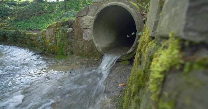 Small water stream running out of an industrial-size concrete pipe in a forest in Europe. Daytime, real time, no people, wide angle