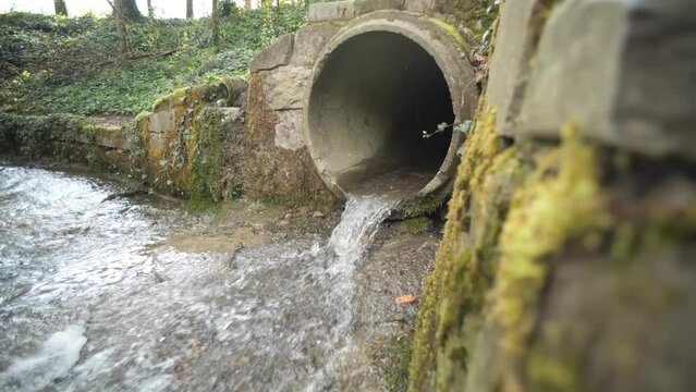 Small water stream running out of an industrial-size concrete pipe in a forest in Europe. Daytime, slow motion, no people, wide angle