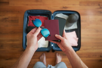 Packing suitcase and planning travel summer adventure. Man holding in hand smart phone, passport and sunglasses above open baggage..