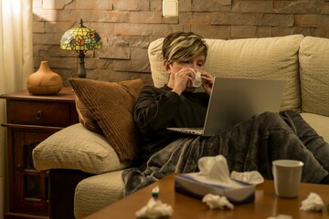 Woman looking at her laptop, while laying on the sofa with high temperature - obrazy, fototapety, plakaty