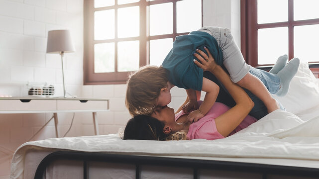 Loving mom playing and bonding with cheerful kid in the morning. Caucasian mother lifting his cute little son in the air to pretend to fly like a plane or superhero with arms out on a bed at home.