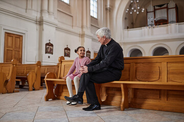 Senior priest talking to African American little girl while they sitting on bench in old church