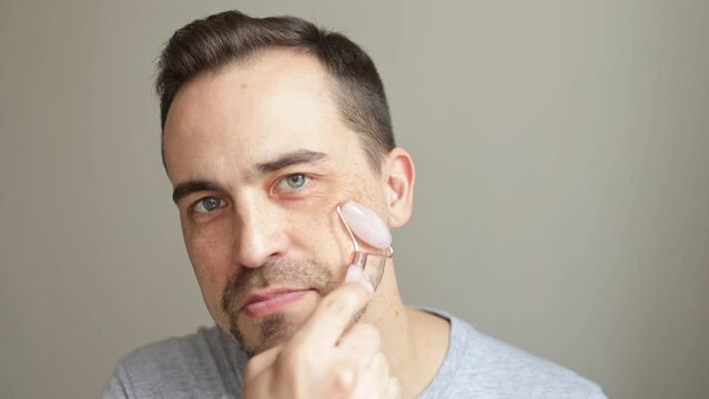 A man massages his face with a jade roller at home looking at the camera.