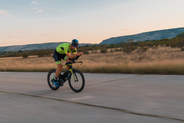Naklejka premium Triathlete riding his bicycle during sunset, preparing for a marathon. The warm colors of the sky provide a beautiful backdrop for his determined and focused effort.