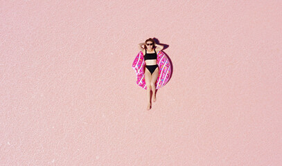 World Tourism Day. Above view of young happy woman in sunglasses and black bikini sunbathing lying on an inflatable pink donut in a sunny pink lake. Enjoying holiday vacations. Place for advertising
