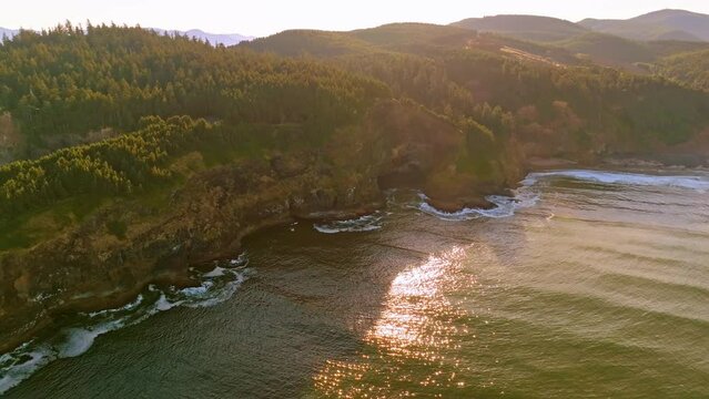 Morning light reveals cliffs, Cape Meares Lighthouse, and Oregon coast; warm glow on rocks, crashing waves, and verdant forest in breathtaking aerial view.