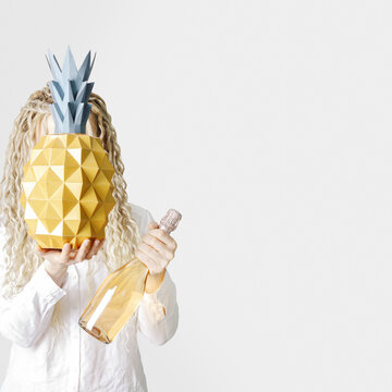 Creative summer festive visual. Woman holding paper pineapple in front of face and rose wine bottle in hand, no face trend concept. Stylish girl with blonde long hair, summer minimal lifestyle