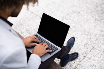 Unrecognizable black businessman surfing the web on laptop with blank screen, sitting outdoors, mockup