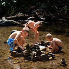 Children happy among nature playing in water of river in summer hot day, building stone pyramid of pebbles. Summer holiday, vacation, free time activity outdor, fun and joy