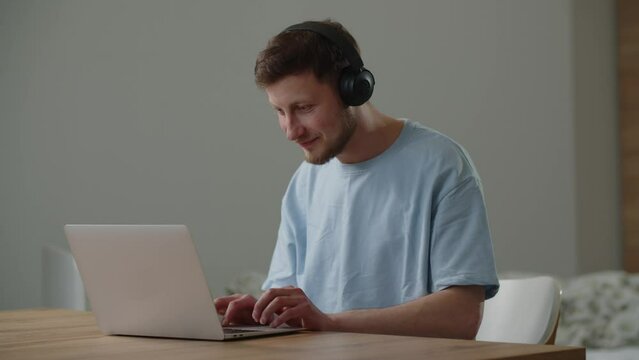 A male freelancer listens to music on headphones while working at a computer. A man in headphones sits at a table and types on a laptop, periodically scrolling through the touchpad.