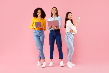 Fototapeta premium Three diverse ladies using different gadgets, networking online, standing on pink background, full length
