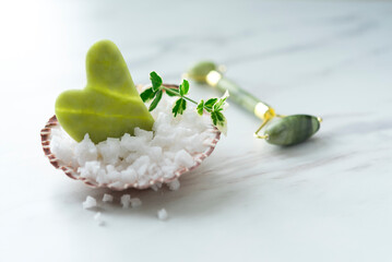 Green jade Gua sha in the sea shell full of sea salt and facial roller on the light marble background. Copy space. Top view