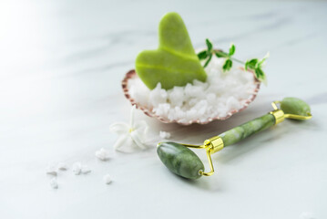 Beautiful spa composition with green jade Gua sha in the big sea shell full of sea salt, facial jade roller, small green branch on the light marble background. Copy space. Flat lay. Freshness 