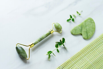 Spa products with green jade facial Gua sha and roller, two small green branches on the light marble background and green bamboo mat in the lower right corner. Copy space. Flat lay