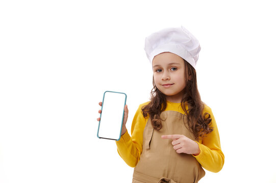 Adorable little chef confectioner, lovely baby girl wearing chef's hat and apron, points at white blank screen of smartphone, smiling looking at camera, isolated background. Copy space for mobile apps - Powered by Adobe