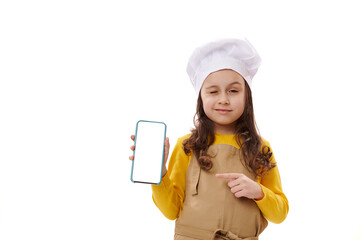 Beautiful Caucasian little girl wearing chef's hat and apron, points at white blank digital screen of smartphone, winking and smiling looking at camera, isolated background. Ordering food. Mobile apps