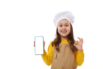 Positive 5-6 years little kid girl wearing chef hat and apron, showing smartphone with blank white screen with free space for insert your mobile apps or advertisement, gesturing with OK sign at camera