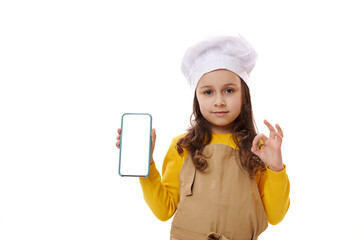 Little girl dressed as chef confectioner, gestures with OK sign and shows at camera a smartphone with blank white digital screen with copy space for mobile apps or advertisement, isolated on white