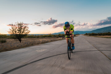Naklejka premium Triathlete riding his bicycle during sunset, preparing for a marathon. The warm colors of the sky provide a beautiful backdrop for his determined and focused effort.