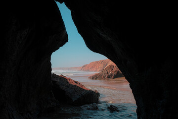 View of Aftas Beach from inside a cave. Waves of the Atlantic Ocean, sand, and rugged rock formations outside. Chill coastal town in Mirleft, Morocco. Landmark popular with surfers, locals, tourists.