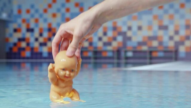 Hand of woman dips small baby doll into clean blue water. Professional shooting of toy underwater in swimming pool close view