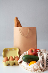 Food in a string bag, eggs in a box and a paper bag of bread are on the table on a grey background. The theme of eco-friendly consumption. Copy space.