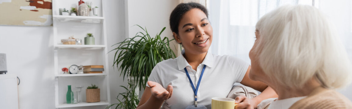 happy multiracial social worker chatting with senior woman while having tea in living room, banner.