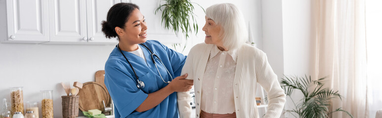 happy multiracial nurse in blue uniform looking at senior woman with grey hair, banner.