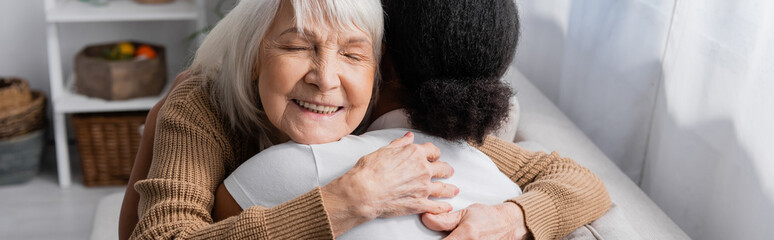 happy senior woman with closed eyes hugging multiracial social worker in living room, banner.