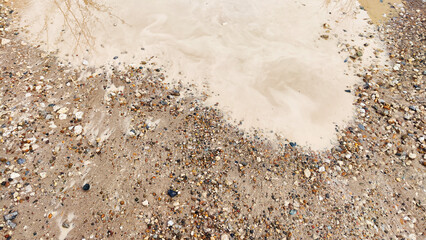 Small dirty brown puddle and small pebbles on the shore. Abstract natural texture, background and copy space. The concept of bad weather in autumn or spring