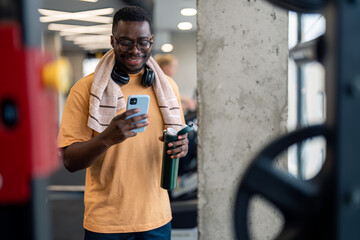 Handsome African American sportsman using mobile phone while taking a break from exercising,...