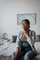 Stressed multiracial woman touching sore throat while sitting on bed at home.