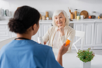 retired woman with grey hair looking at multiracial nurse with medication on blurred foreground.