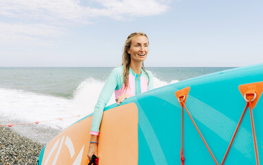 A young woman carries a SUP board to the sea and smiles.