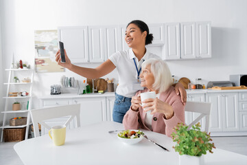 happy multiracial social worker taking selfie with retired woman during lunch in kitchen.
