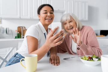 happy multiracial social worker holding smartphone while senior woman waving hand during video call.