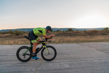 Fototapeta premium Triathlete riding his bicycle during sunset, preparing for a marathon. The warm colors of the sky provide a beautiful backdrop for his determined and focused effort.