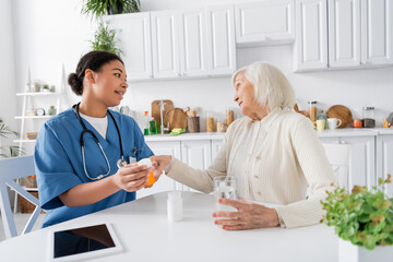 Fototapeta na wymiar multiracial nurse showing medication to skeptical senior woman next to digital tablet on table.