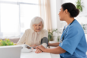 joyful multiracial nurse measuring blood pressure of senior woman with grey hair.