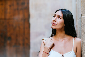 Dreamy Latin American woman adjusting backpack on shoulder and admiring street while leaning on...