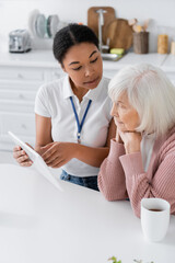 positive multiracial social worker holding digital tablet near senior woman in kitchen.
