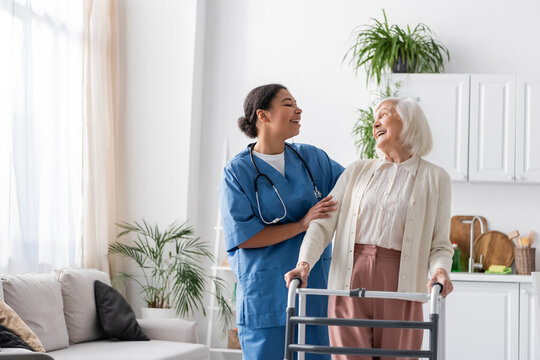 Happy Multiracial Nurse In Uniform Laughing With Retired Woman While Standing Near Walking Frame At Home.