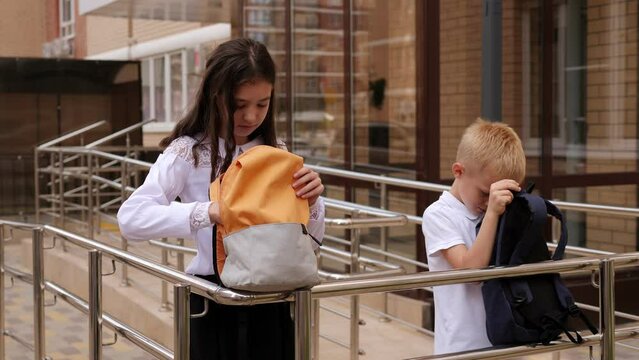 Close-up of a boy and a girl in school uniform standing near the school and looking into open backpacks.