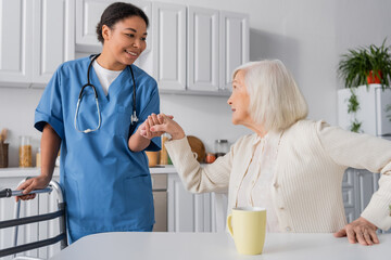 Fototapeta premium happy multiracial nurse in blue uniform holding hand of senior woman with grey hair while standing near walker.